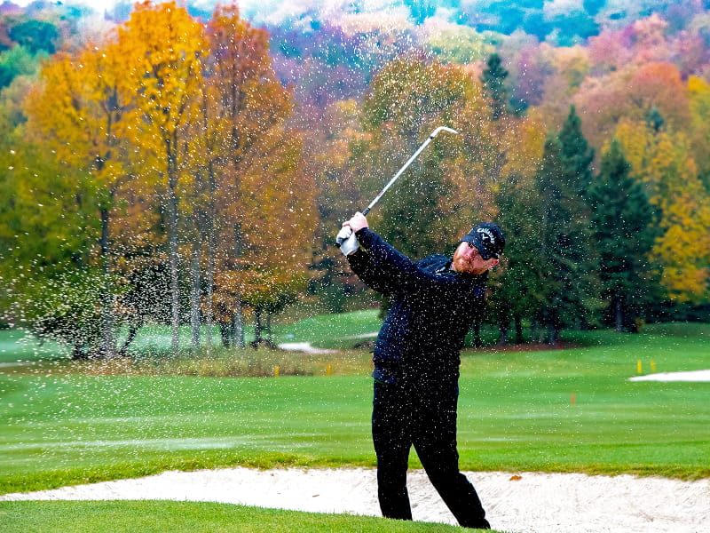 Golfer wearing dark clothing hits from a sand bunker as sand sprays into the air on a fall-colored golf course backdrop.