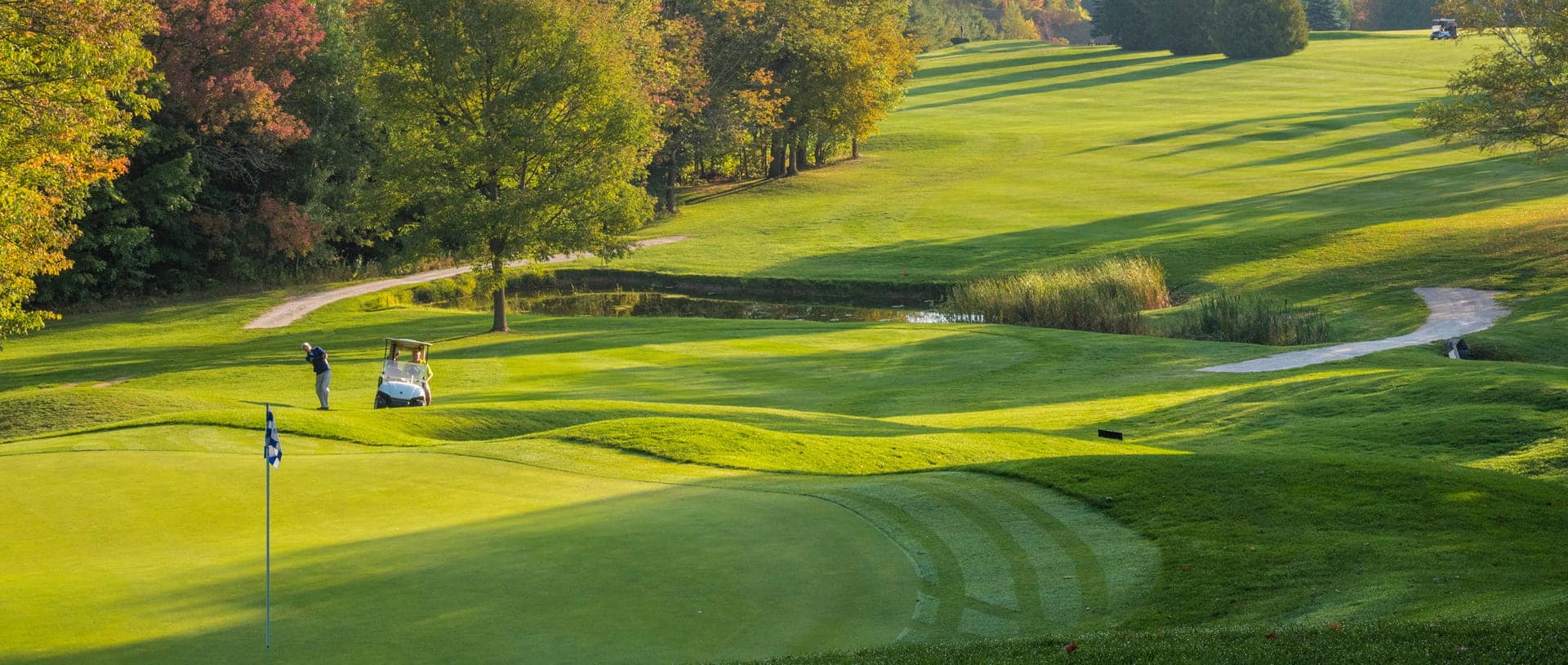 Golfer swinging on a sunny, rolling golf course with a nearby cart and green flag in the foreground.