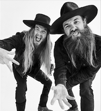 Two bearded men wearing black hats and dark clothing lean toward the camera with outstretched hands, smiling playfully against a white background.