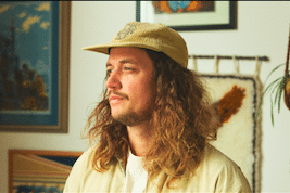 Portrait of a man with long wavy hair wearing a tan hat and light shirt, indoors near framed artwork on the wall.