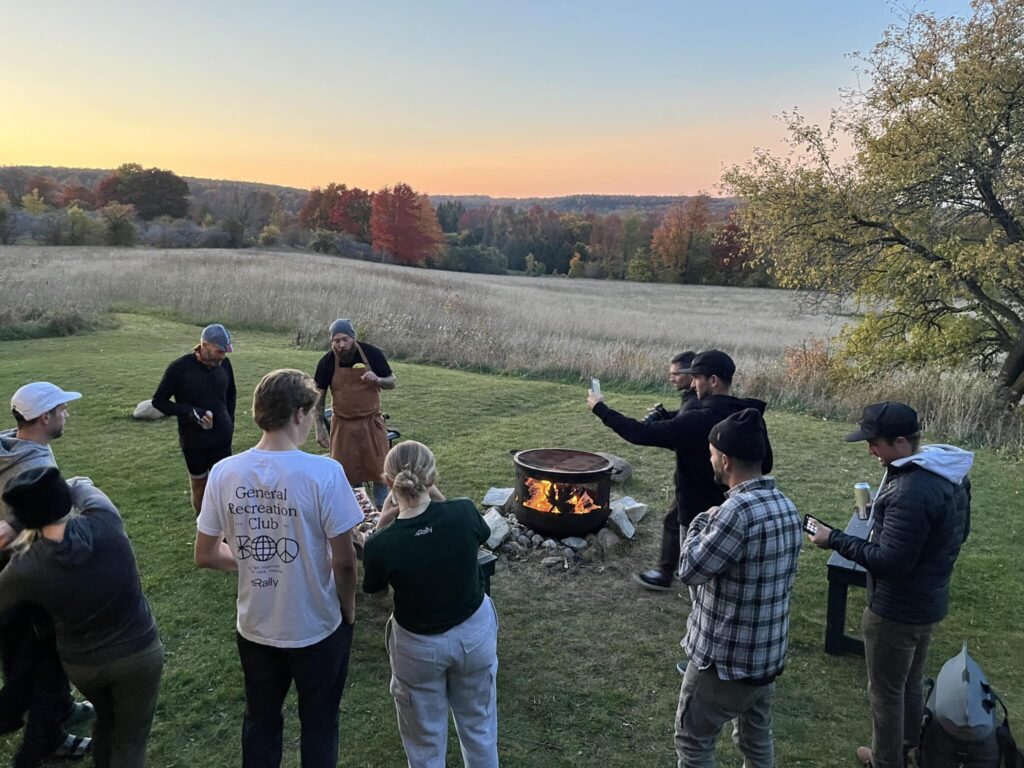Group of people standing and sitting around a lit fire pit in a grassy field at sunset, with autumn trees in the background and a clear sky.