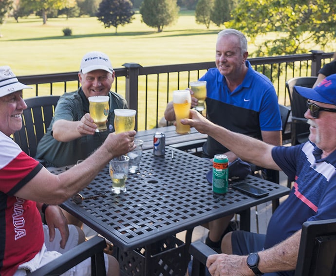 Five men sit around a metal patio table on a sunny golf course, raising beer glasses in a toast.