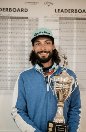 Smiling man in a blue pullover and a turquoise TaylorMade cap holds a large silver trophy, with leaderboard score sheets in the background.